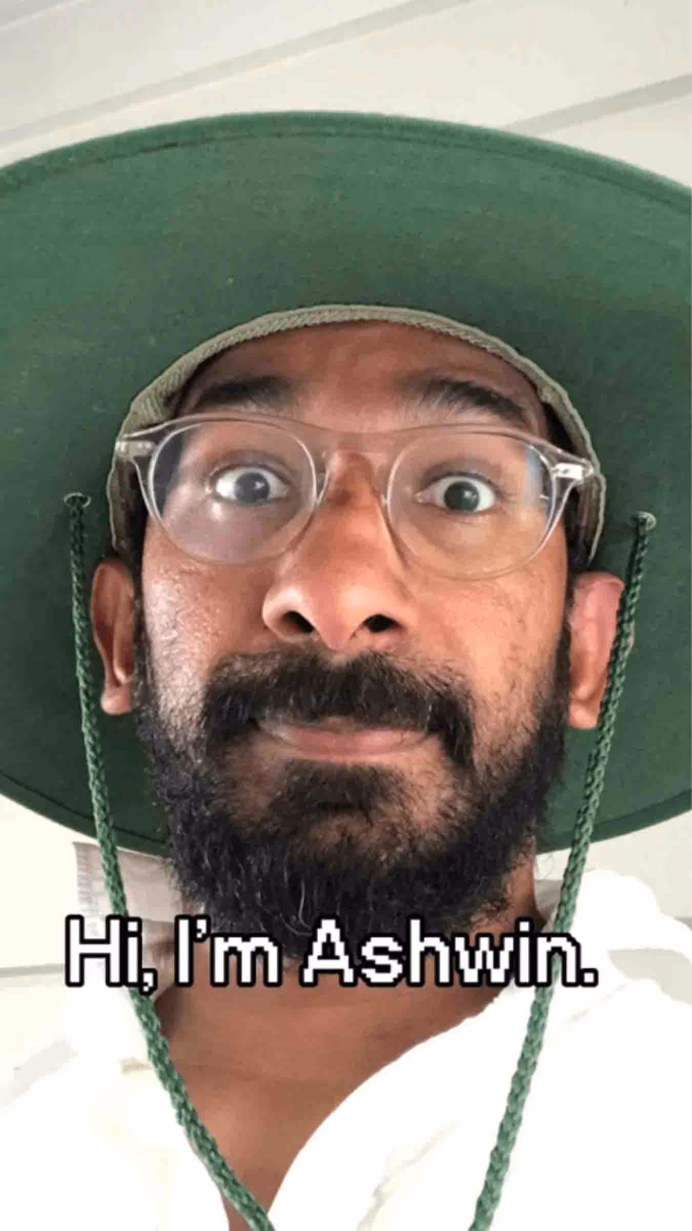 A close-up selfie of a man wearing glasses and a wide-brimmed green hat, with on-screen text that reads 'Hi, I'm Ashwin.