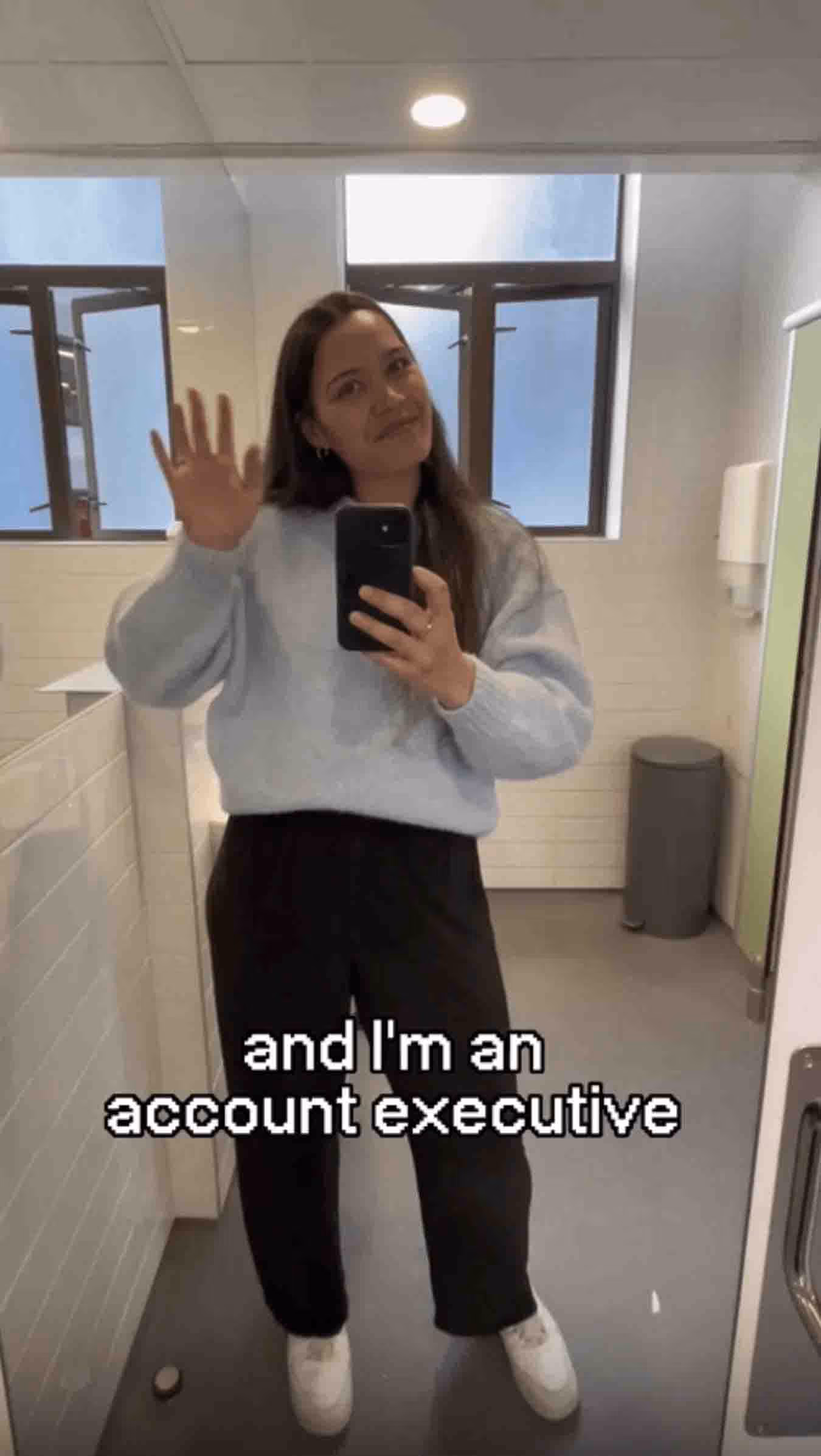 A young woman waving at the mirror while taking a selfie, with on-screen text that reads 'and I'm an account executive'.