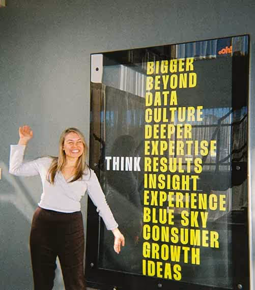 A woman smiling and posing next to a bold agency poster that reads 'Think Bigger, Beyond, Data, Culture…
