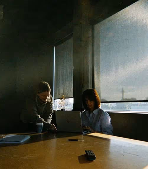 Two women working together at a laptop in a softly lit meeting room, focused on a project in progress.