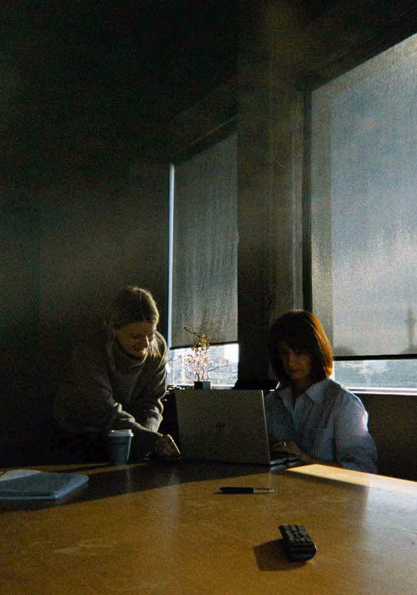 Two women working together at a laptop in a softly lit meeting room, focused on a project in progress.