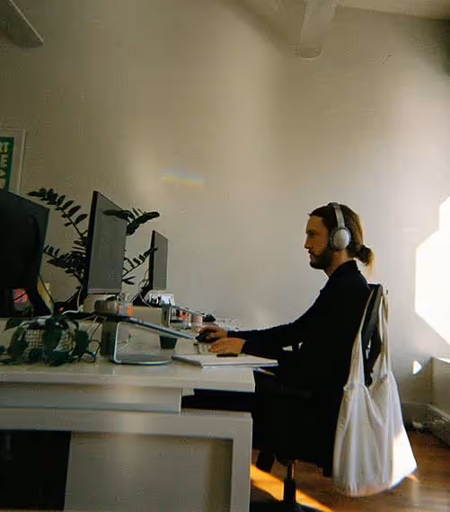 A man wearing headphones works at a dual-monitor setup in a bright, plant-filled office.