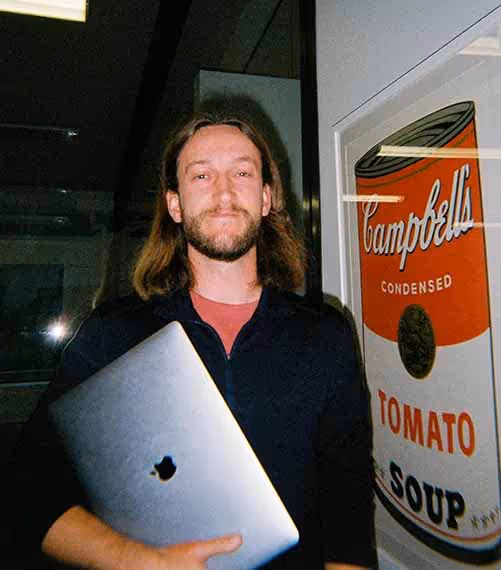 A man with long hair smiles while holding a laptop next to a framed Campbell’s soup poster in a creative office.