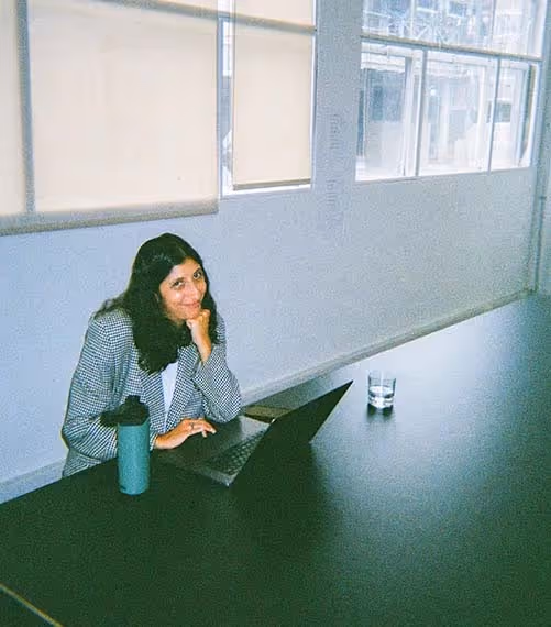 A woman smiling at a long meeting table with her laptop and coffee, mid-workday in a light-filled office.