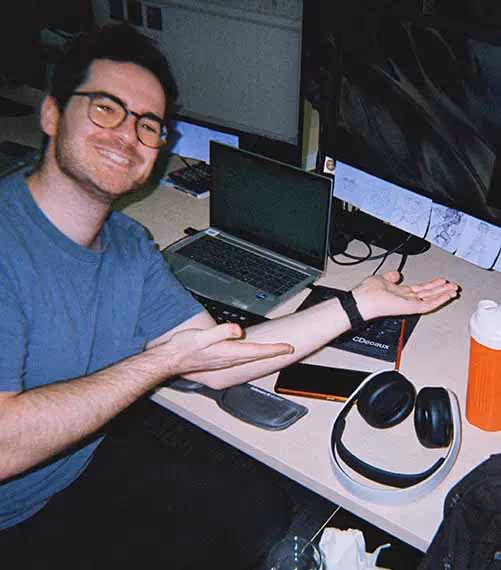 A man smiling and gesturing enthusiastically at his workspace, surrounded by monitors and tech gear in a media-focused advertising role.