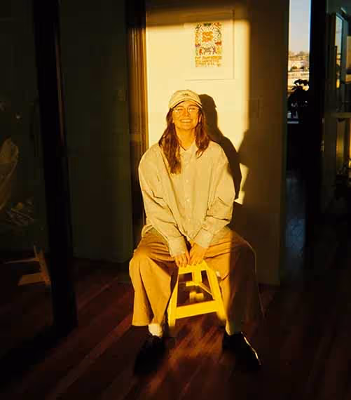 A person sitting on a stool in golden-hour light, smiling in the hallway of a creative workspace.