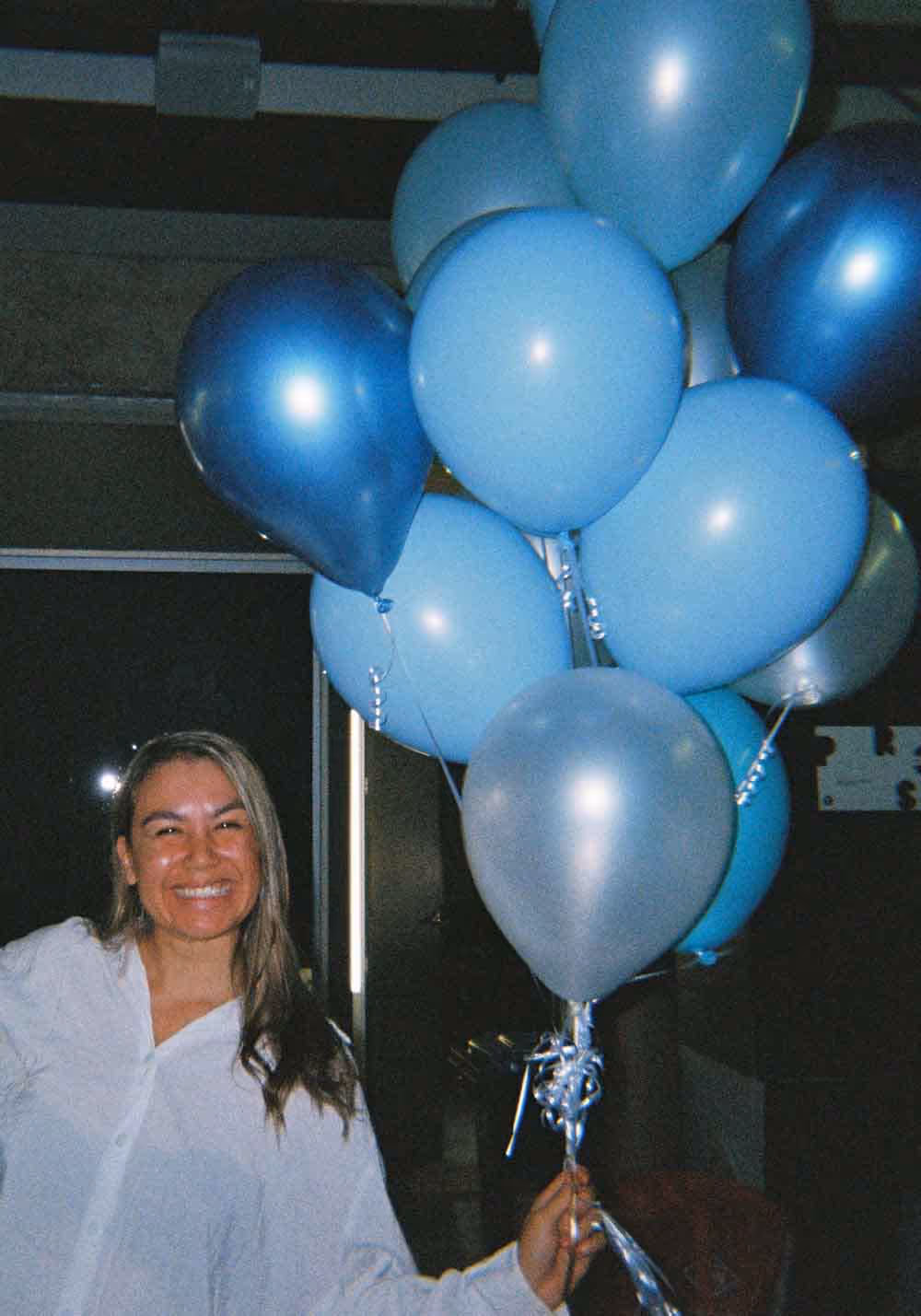 A woman smiling and holding a bunch of blue balloons at an agency event, showing the celebratory side of advertising culture.