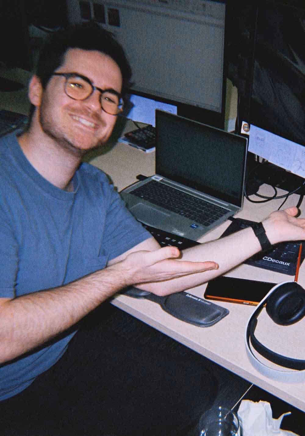 A man smiling and gesturing enthusiastically at his workspace, surrounded by monitors and tech gear in a media-focused advertising role.