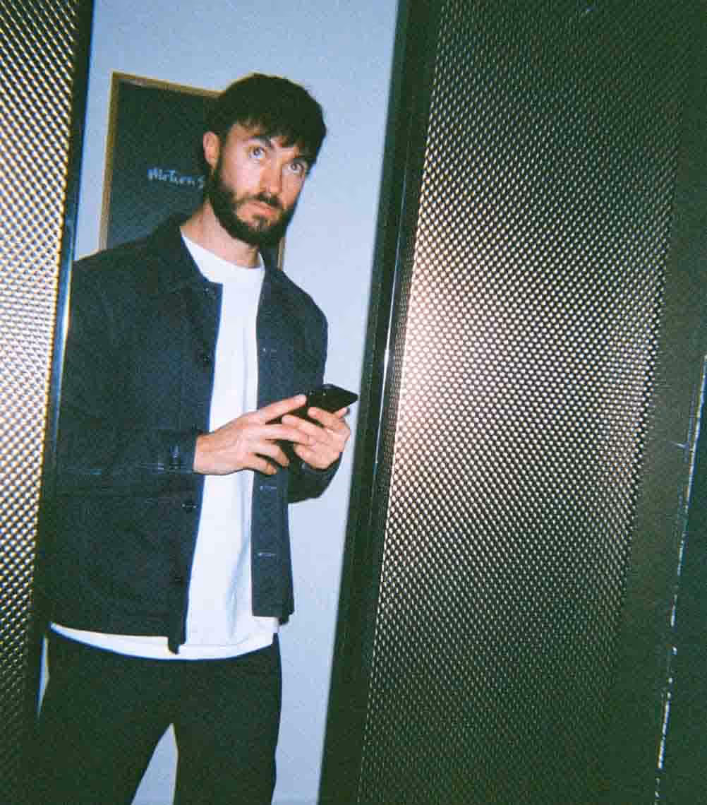 A man standing in a modern office hallway holding a phone, capturing a candid moment from agency life in the advertising industry.