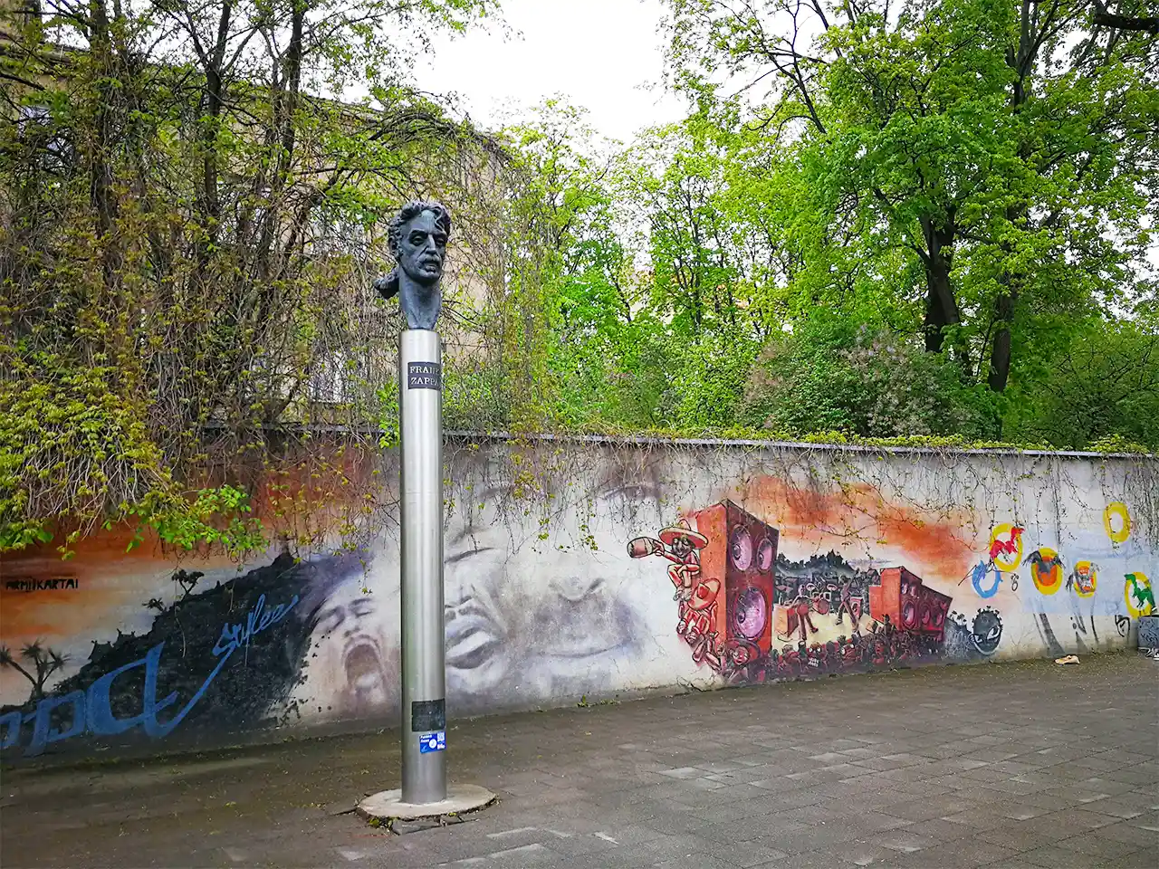 Паметник на Франк Запа в Ужупис на фона на изрисувана плътна ограда и буйна растителност