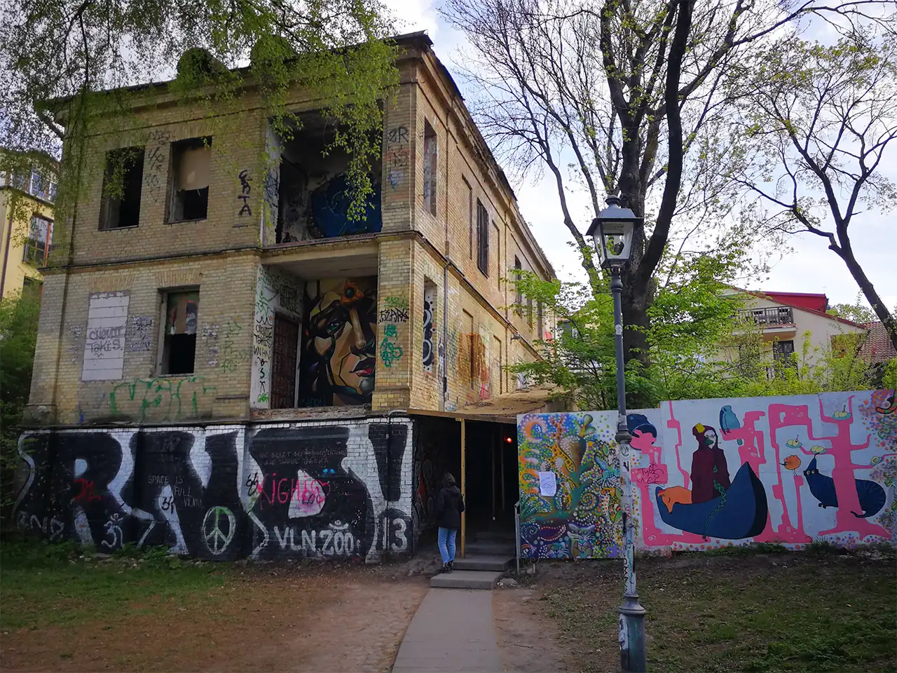 Графити по фасадите и защитната ограда край стара необитаема тухлена сграда с липсващи прозорци в паркова среда