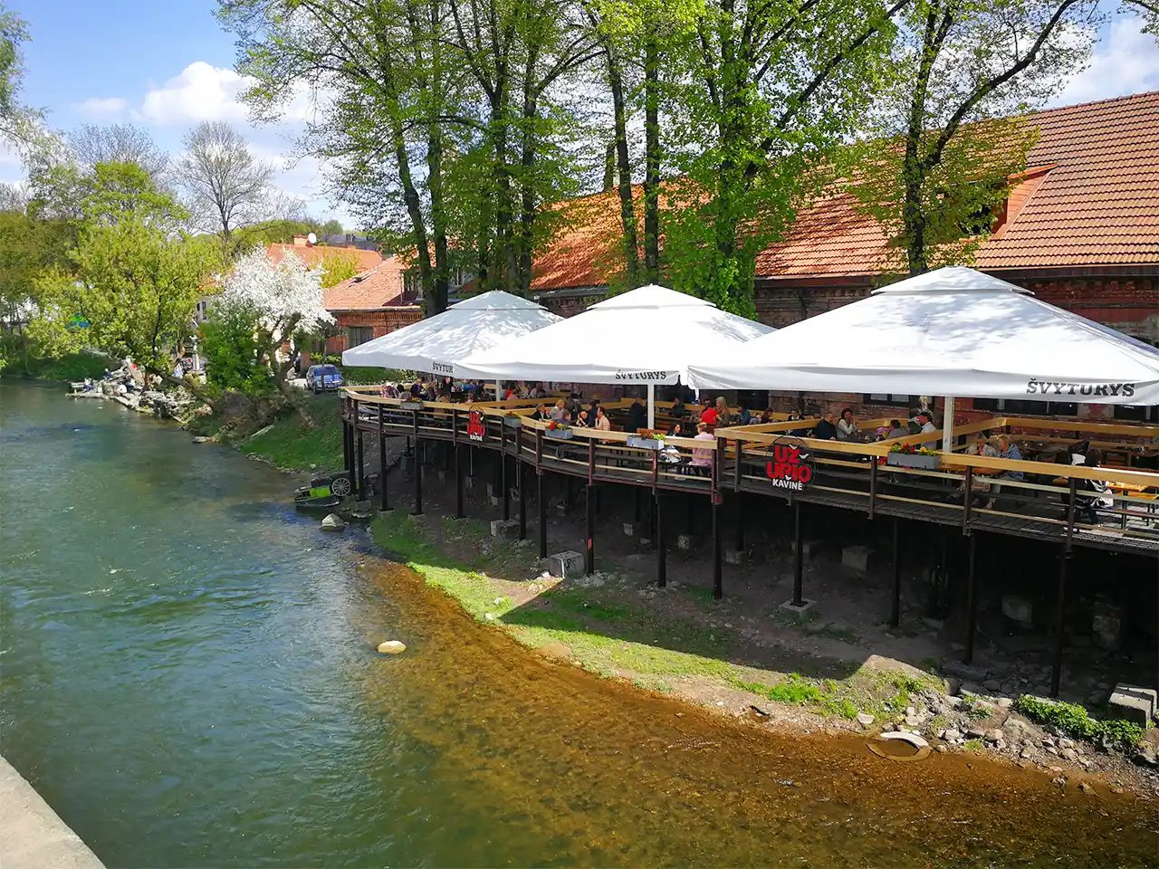 Наддаващи се над граничната за Република Ужупис река Вилня дървени тераси на заведение, изпълнени с разговарящи хора под сянката на големи бели чадъри