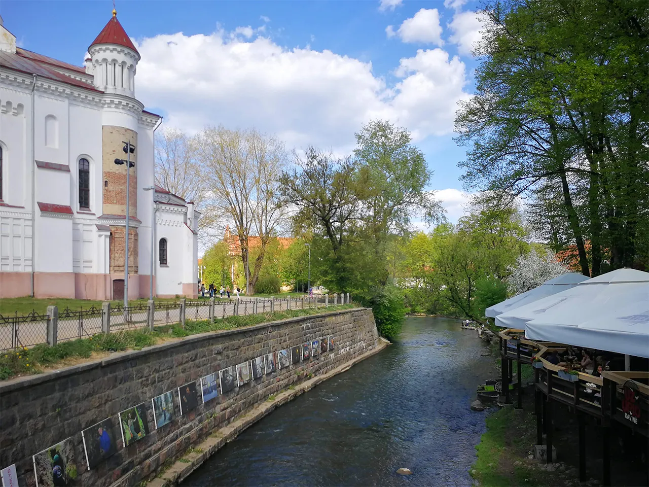 Непринудено аранжирана фотоизложба и дървени тераси на заведение от двете страни на коритото на река Вилня във Вилнюс, сред зеленина и пешеходни алеи