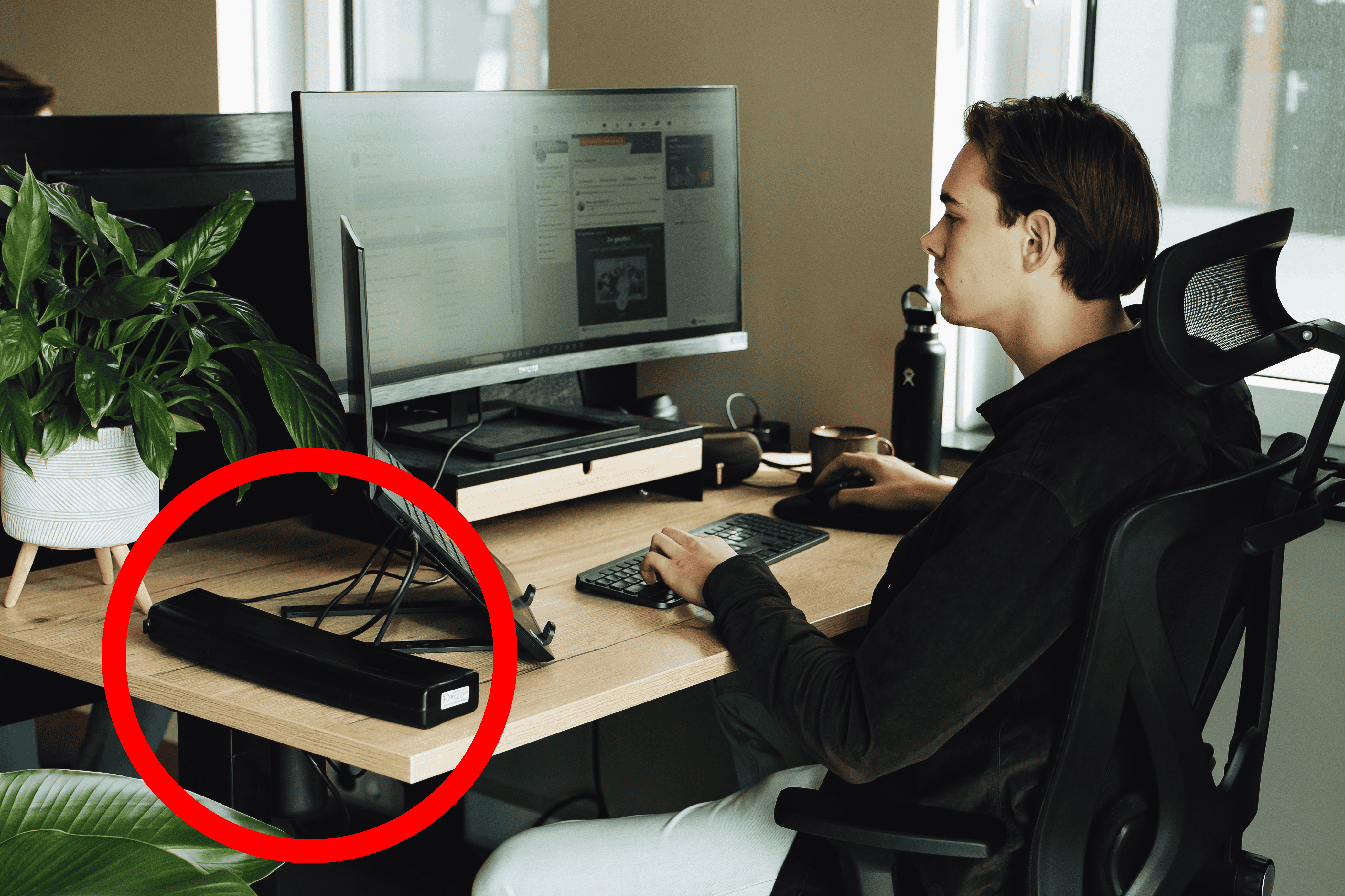 Een vrouw zit aan een bureau bij een groot raam op kantoor en bekijkt documenten terwijl ze aantekeningen maakt. Op het bureau staan een laptop, een waterfles en een stekkerdoos met een e-bike accu die aan het opladen is.