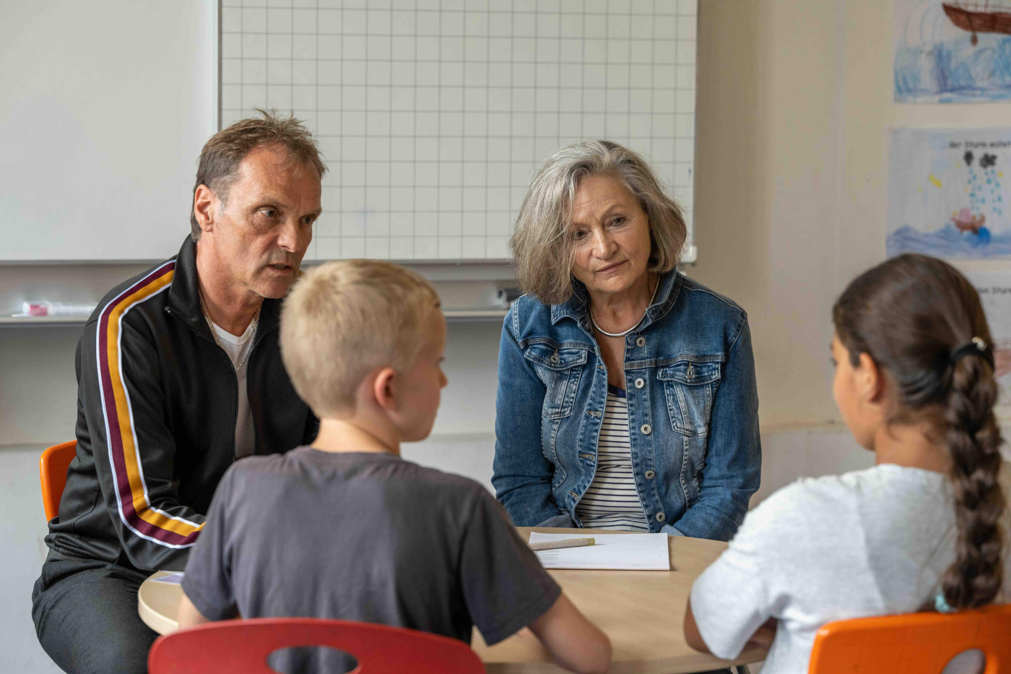 Wir sehen zwei erwachsene Mediatoren, die mit zwei Kindern im zugewandten Gespräch an einem Tisch in der Schule sitzen.