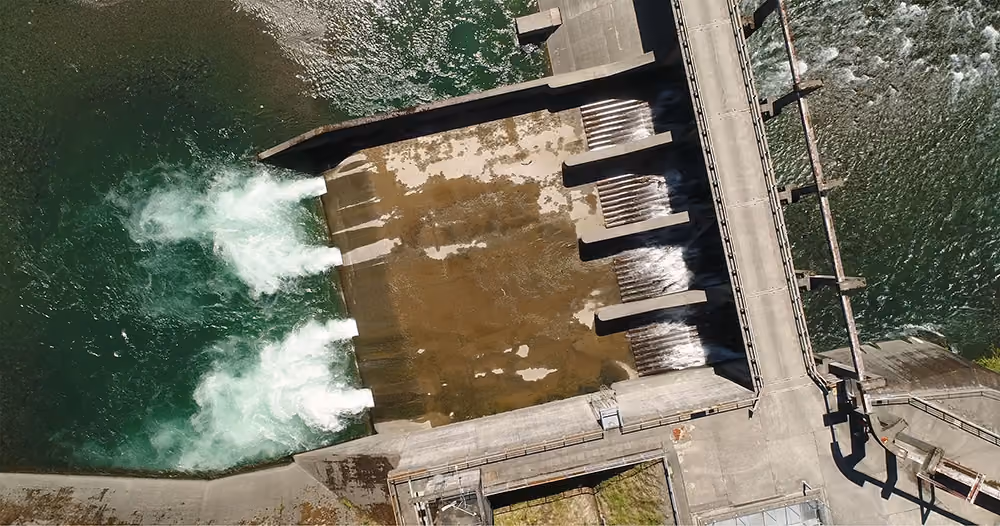 Aerial view of a concrete dam releasing water through multiple gates into a river.