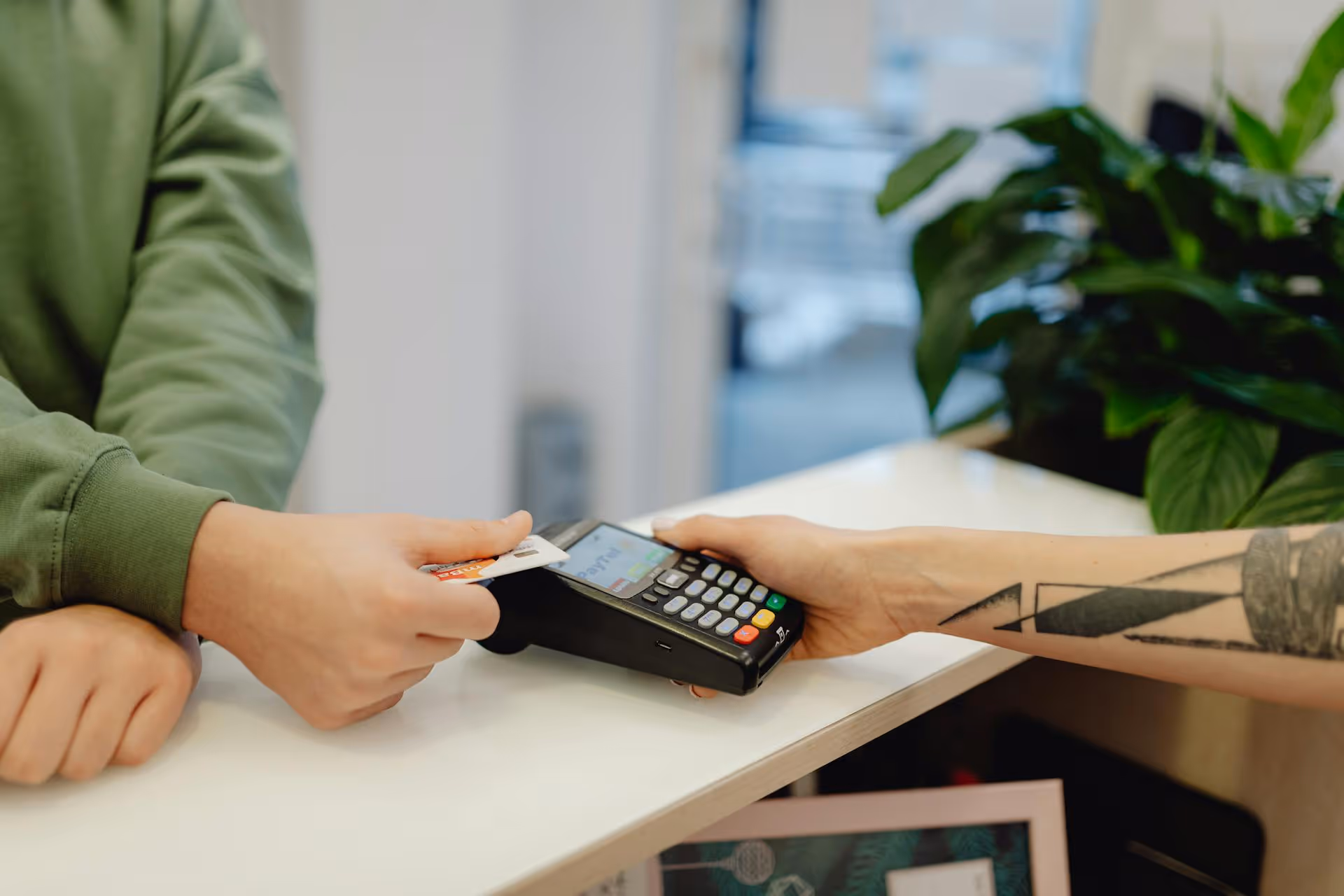 Person holding a credit card near a payment terminal held by another person with a tattooed arm for contactless payment.