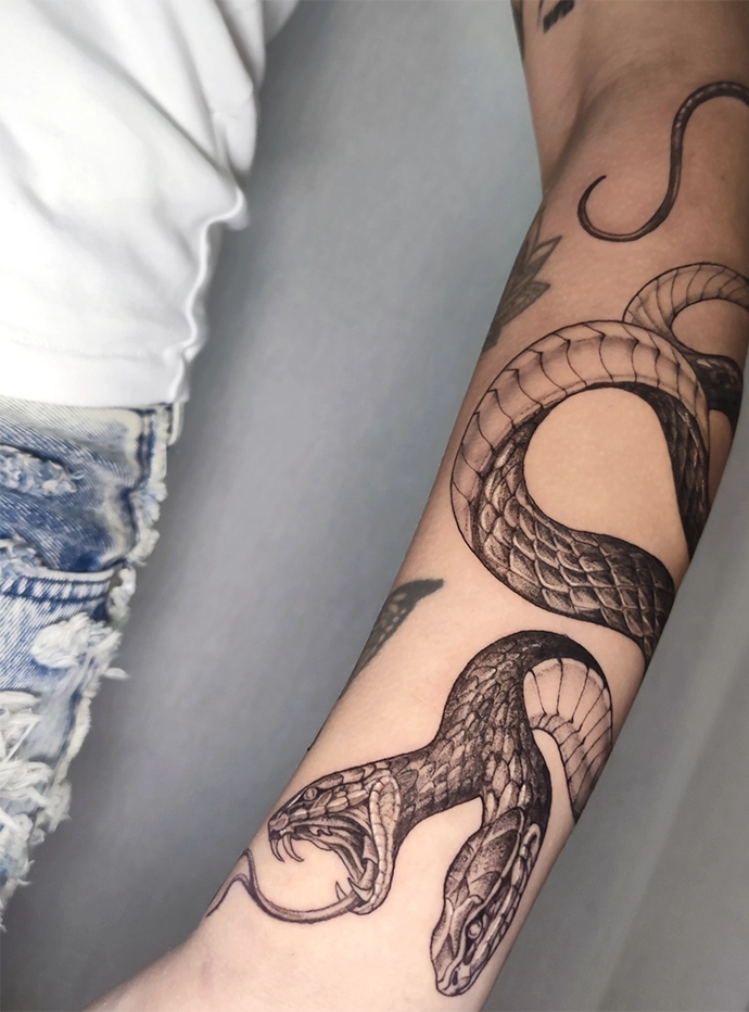 Zwarte tattoo van een slang met twee koppen op de onderarm van een persoon.