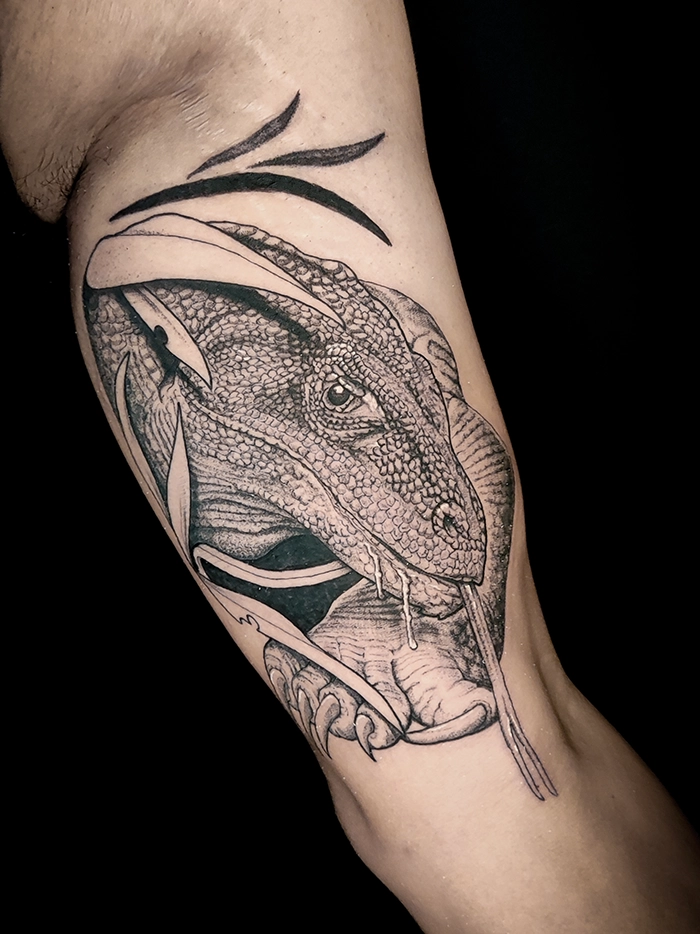 Zwart-wit tattoo van een hagedis met lang uitgestoken tong op een arm.