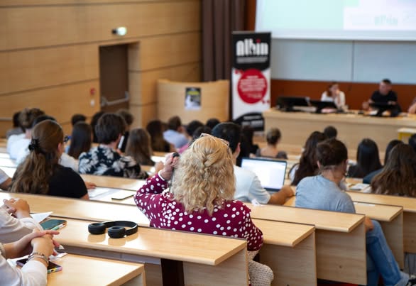 Audience seated in a lecture hall listening to two presenters at a front podium.
