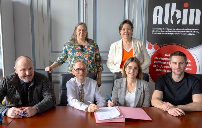 Six people gathered around a table with documents, two standing behind and four sitting, with an Albin organization banner in the background.