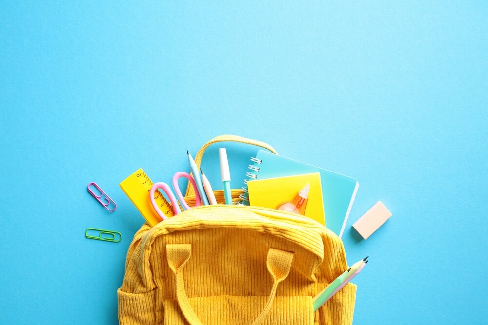 See the Good: Help Us Fill Backpacks for the Boys and Girls Club This Summer!  Back to School Concept. Brightly colored yellow backpack overflowing with essential school supplies such as notebooks, pens, pencils, scissors, and a ruler on a vibrant blue background. Flat lay.