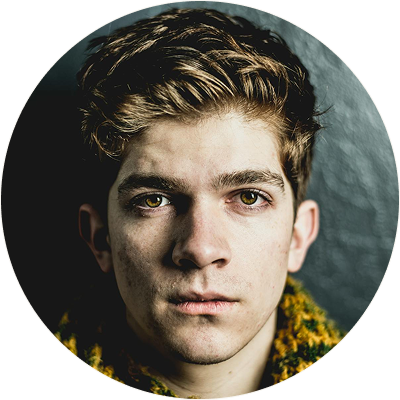 Close-up portrait of a young man with light brown hair and green eyes wearing a yellow and green knitted scarf, looking directly at the camera with a neutral expression.