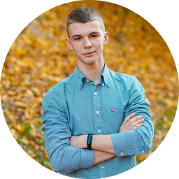 Young man with short hair wearing a blue button-up shirt standing outdoors with arms crossed.