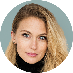 Close-up portrait of a blonde woman with blue-green eyes wearing a black turtleneck against a muted blue background.