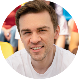 Smiling young man with short brown hair wearing a white shirt seated outdoors with blurred colorful background.