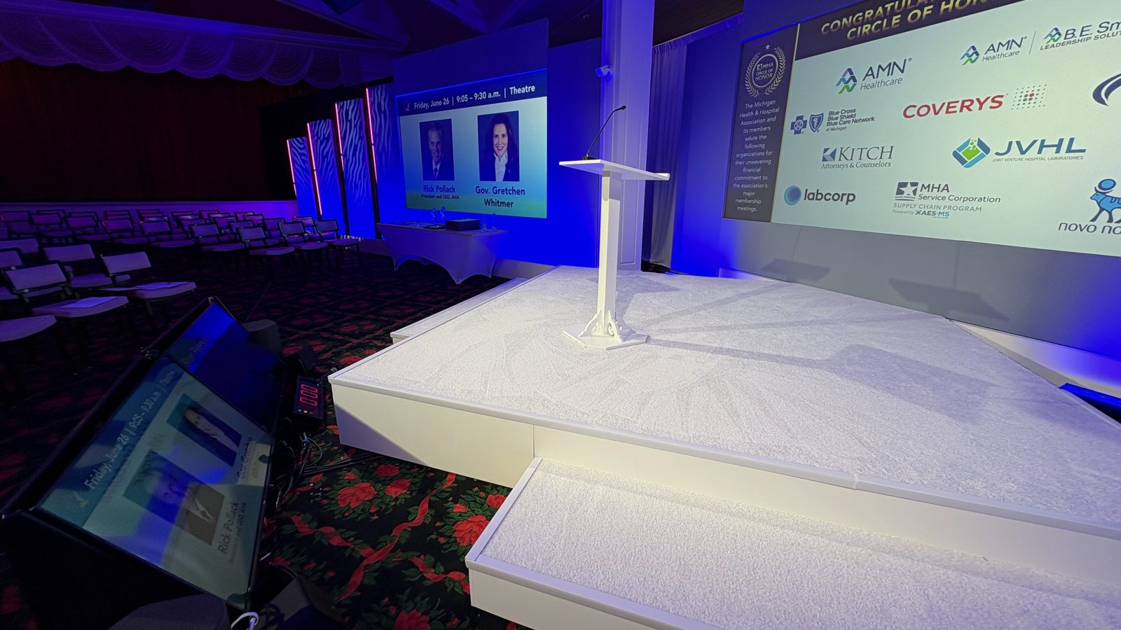 An event stage with a clear podium and a sponsor logo wall in blue lighting