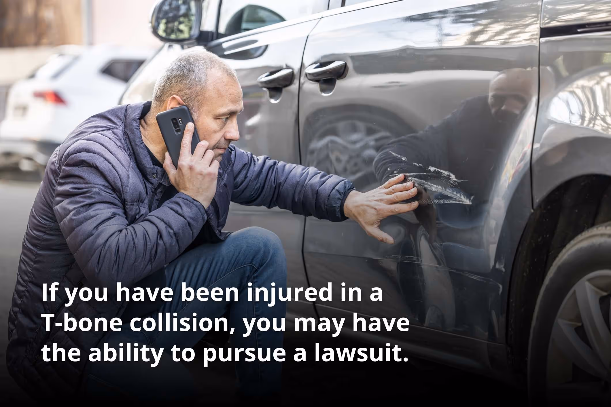 A man kneels down next to his car and calls his insurance company after a collision.