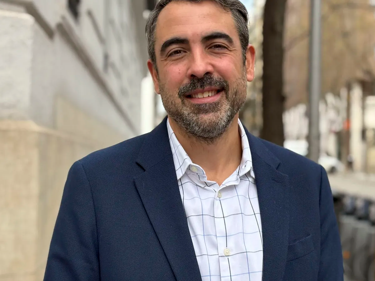 Man smiling outdoors on a city street, wearing a blazer and button down shirt.