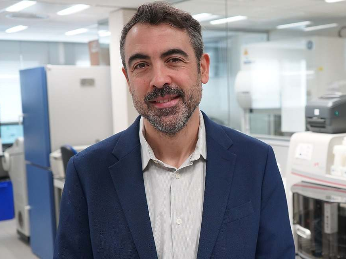 Man standing and smiling in a laboratory environment with scientific equipment in the background.