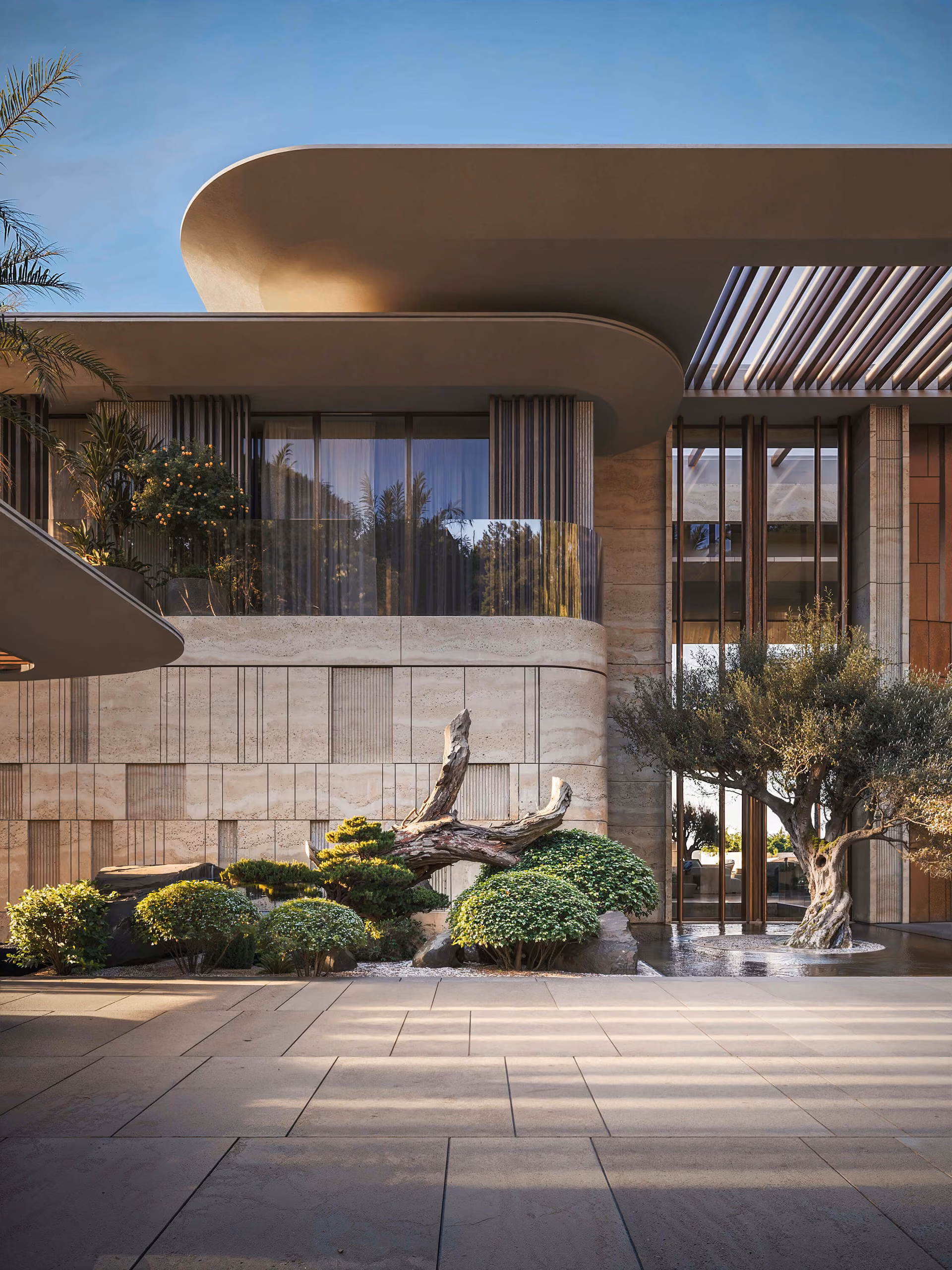 Stone and glass facade detail with a curved roof, ornamental trees and entrance paving.