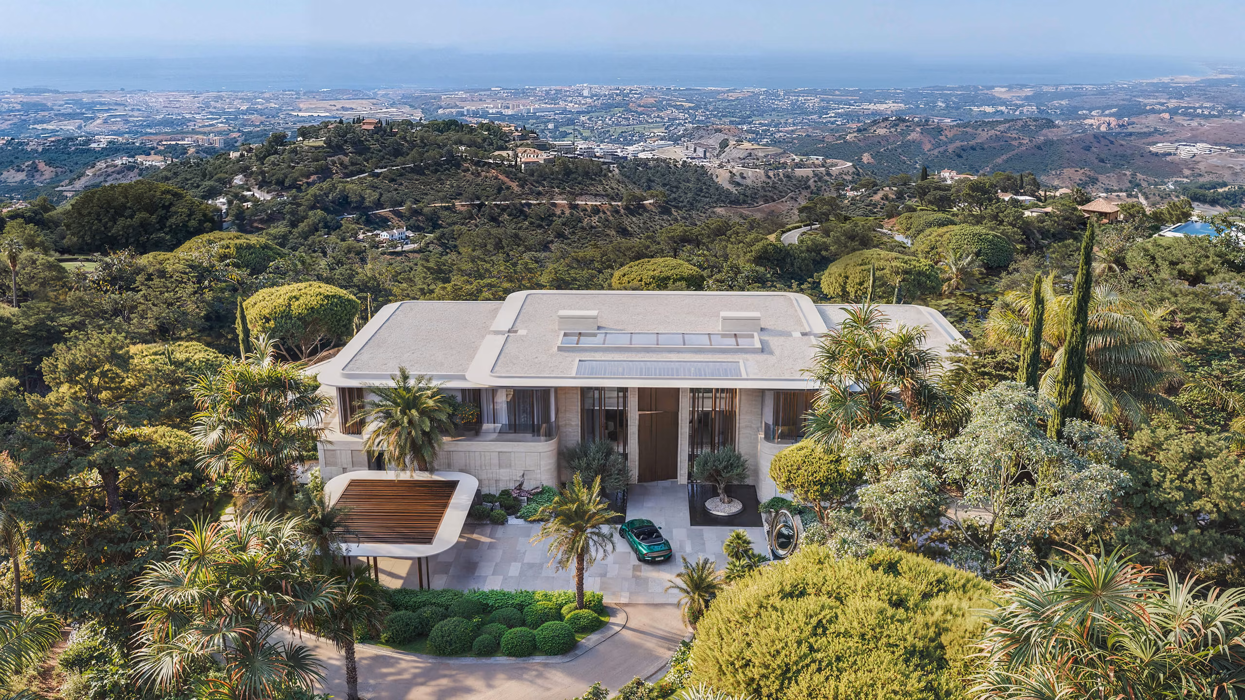 Aerial view of the villa entrance among palms, with the flat roof and coast beyond.