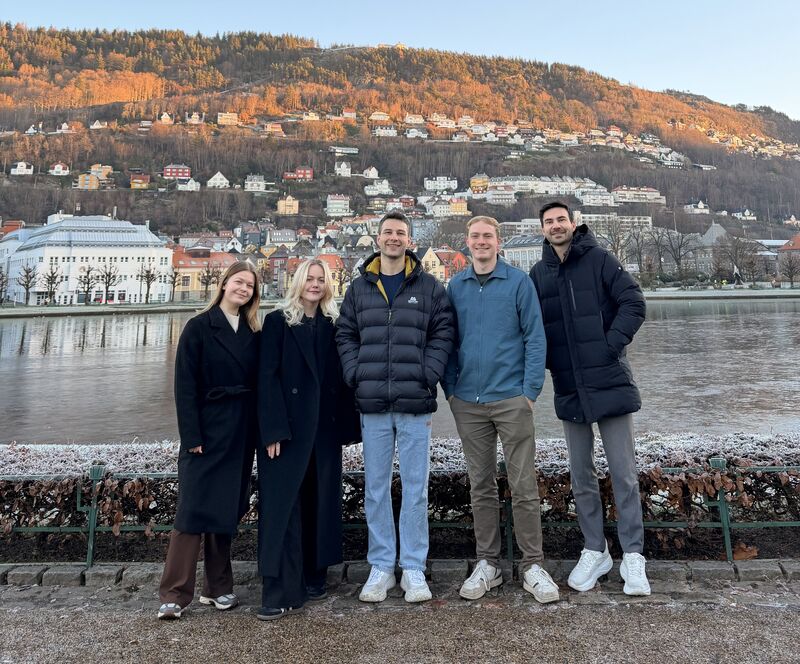 Mann i blå genser smilende foran en innsjø med by og skogkledd ås i bakgrunnen under klar himmel.