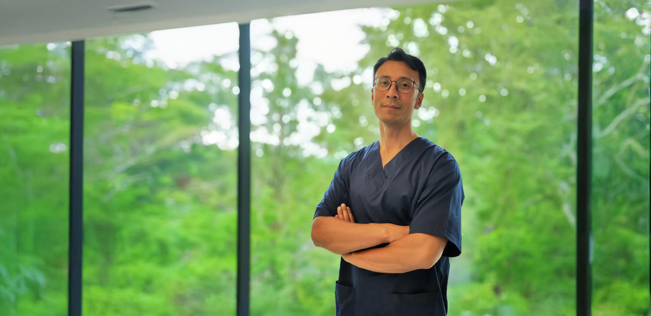 Prof. Dr. med. Thiha Aung in OP-Kleidung vor einer Fensterfront mit Bäumen