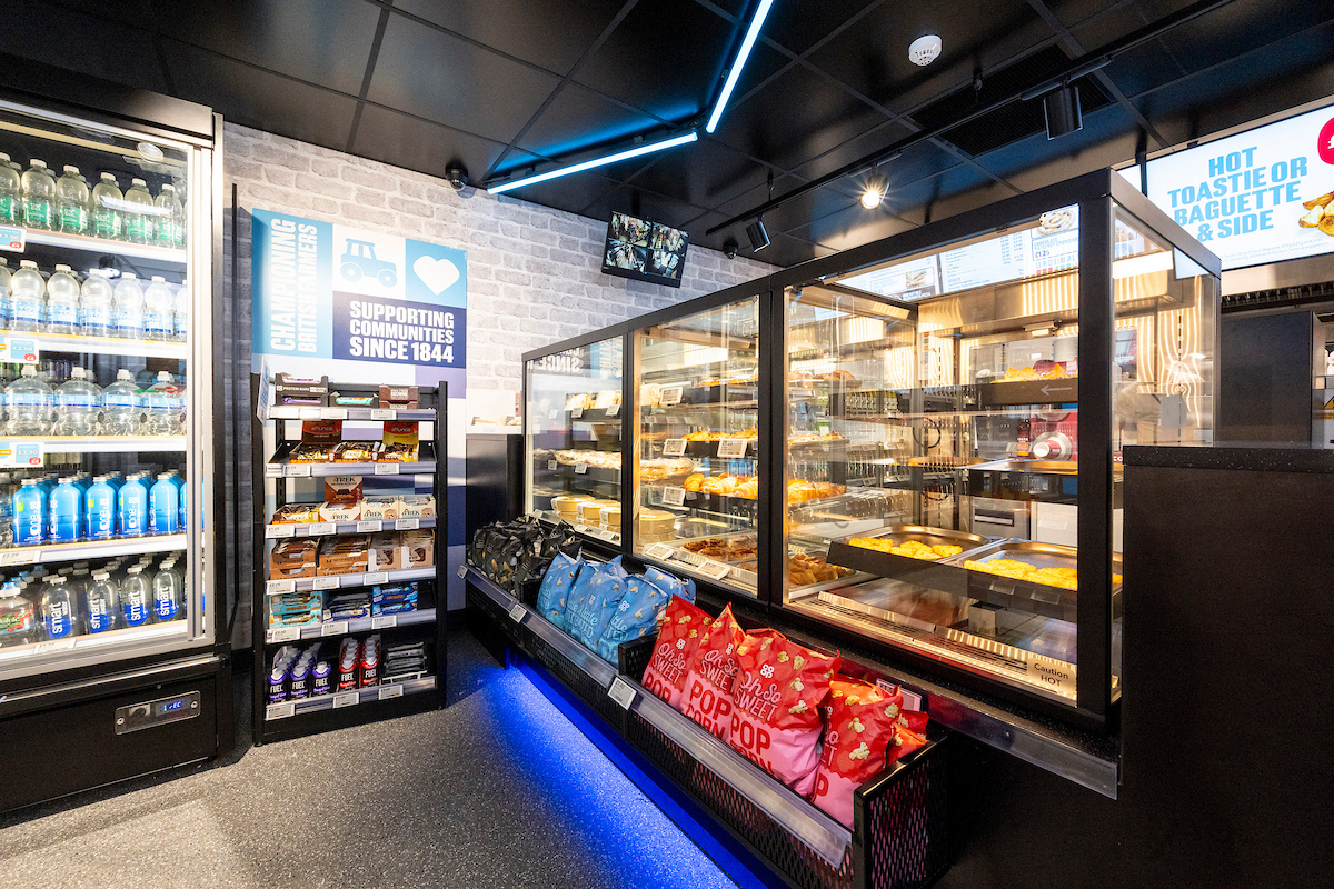 Hot food counter inside Co-op On the Go featuring fresh baked goods, packaged snacks, and bottled drinks under digital signage.