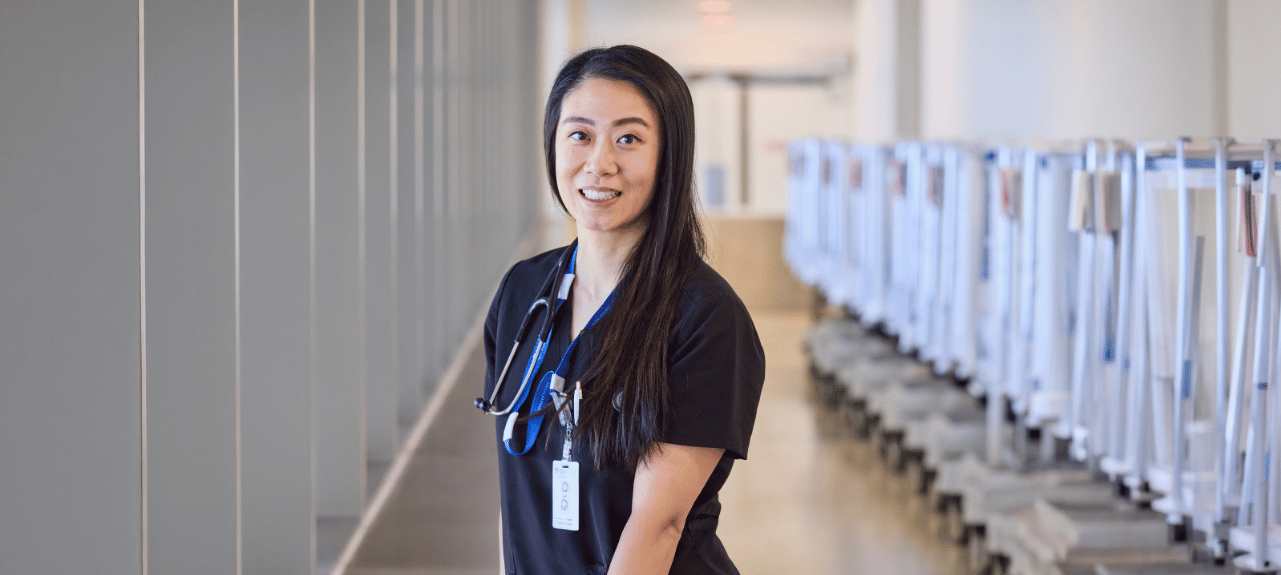 Physician wearing black scrubs and a stethoscope, smiling in a bright hospital corridor beside a row of patient care bays.