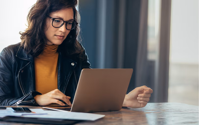 Young woman in glasses and a leather jacket working on a laptop at a wooden table with documents and a phone nearby.