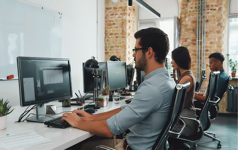 Three diverse professionals working at computers in a modern office with exposed brick walls.