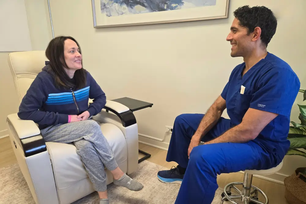 A woman sits in a recliner smiling and talking with a male doctor in blue scrubs seated on a stool in a clinical setting discussing NAD+ and IV ketamine therapy