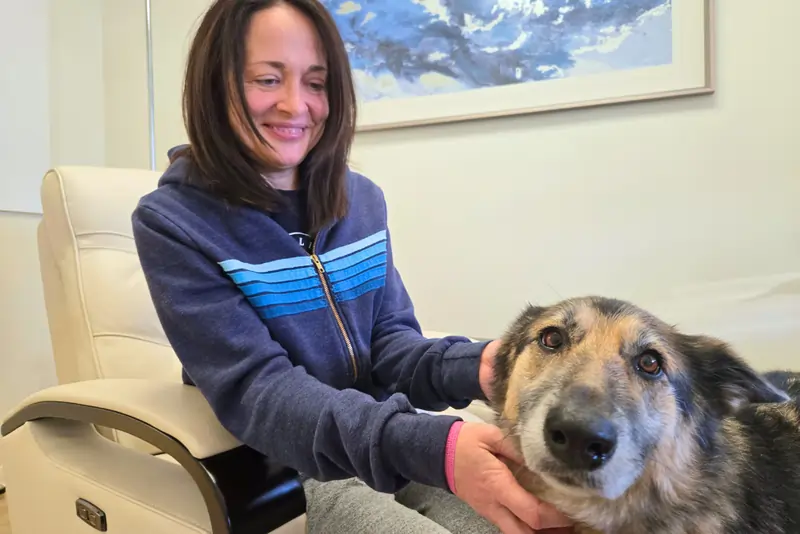 Smiling woman struggling with depression and anxiety sitting in a chair gently holding the face of a large, calm dog with a gray and brown coat.