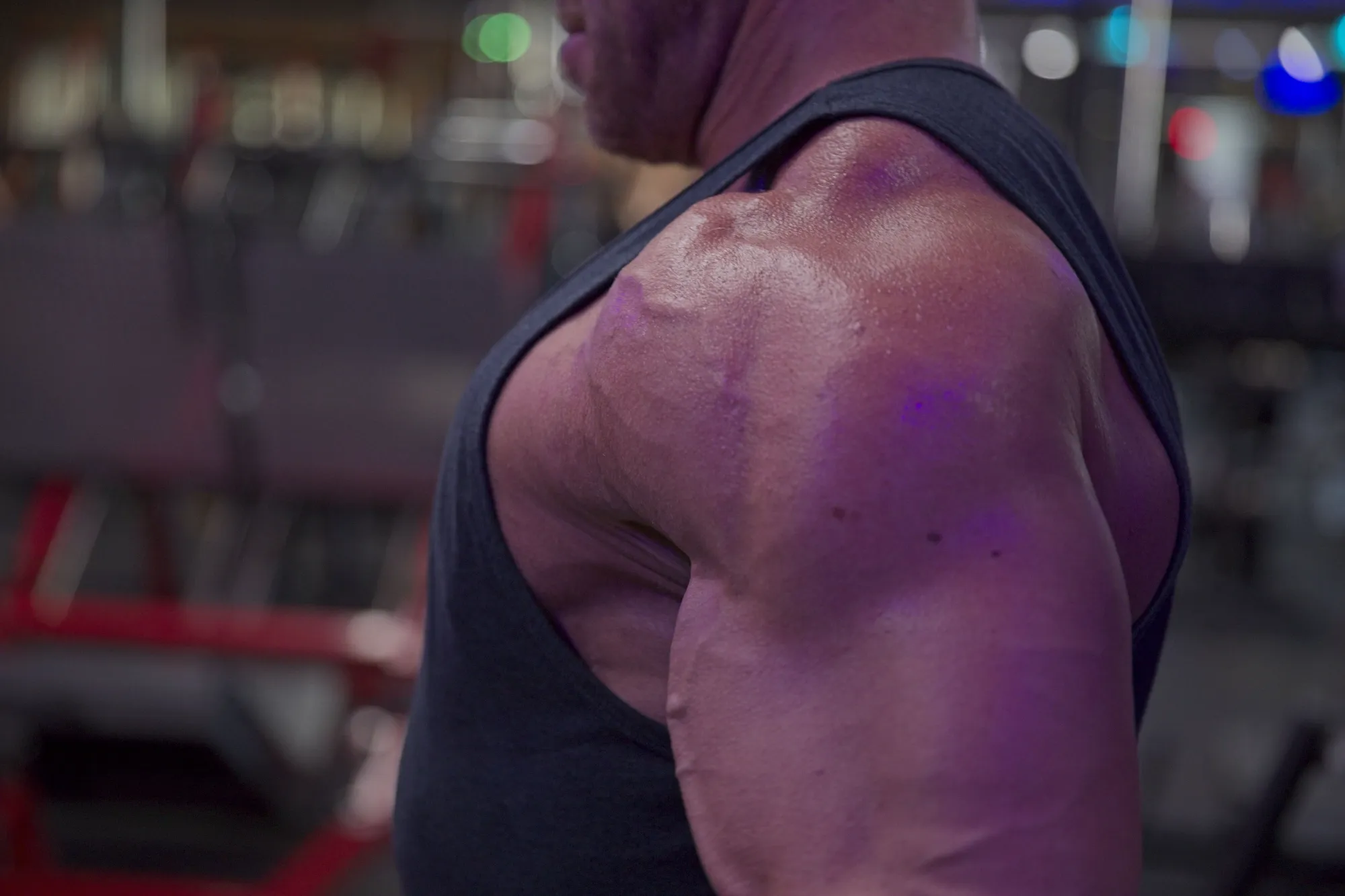 Close-up of a muscular man's shoulder and upper arm wearing a black sleeveless shirt in a gym.