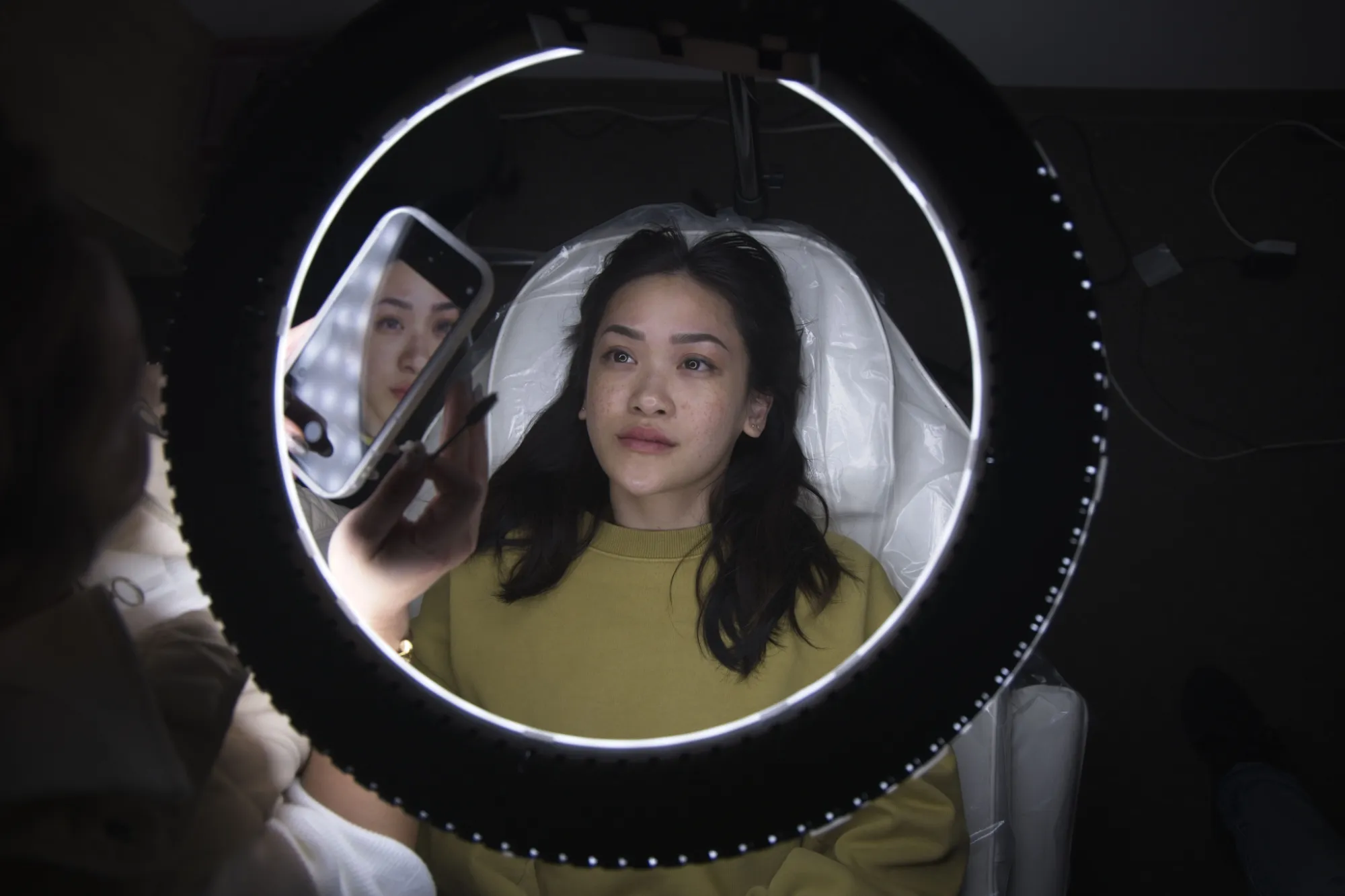 Woman lying on a treatment bed looking at her reflection in a mirror held by another person, illuminated by a circular ring light.