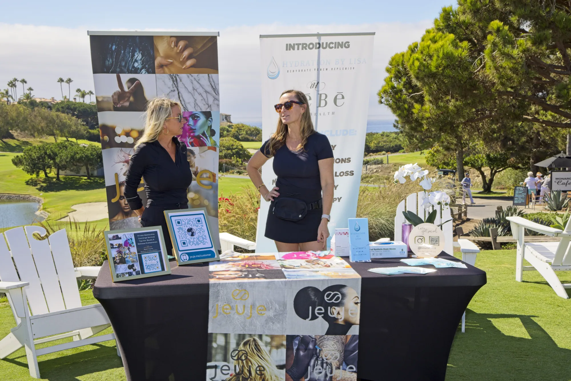 Two women standing behind an outdoor promotional table with Jeuje and Hydration by Lisa banners, set in a sunny park with green grass and trees.