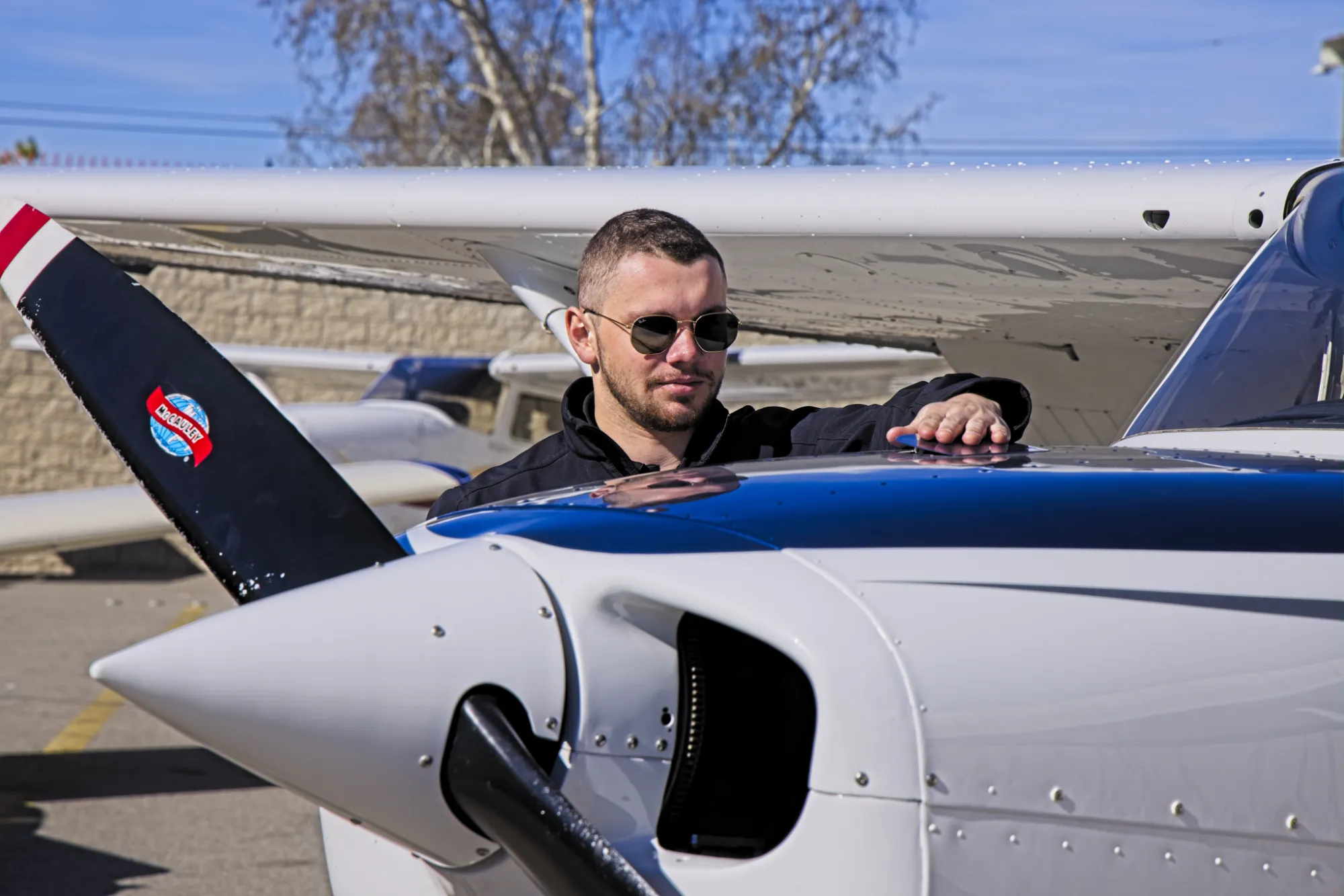 Man wearing sunglasses inspecting the top of a small white and blue airplane near its propeller.
