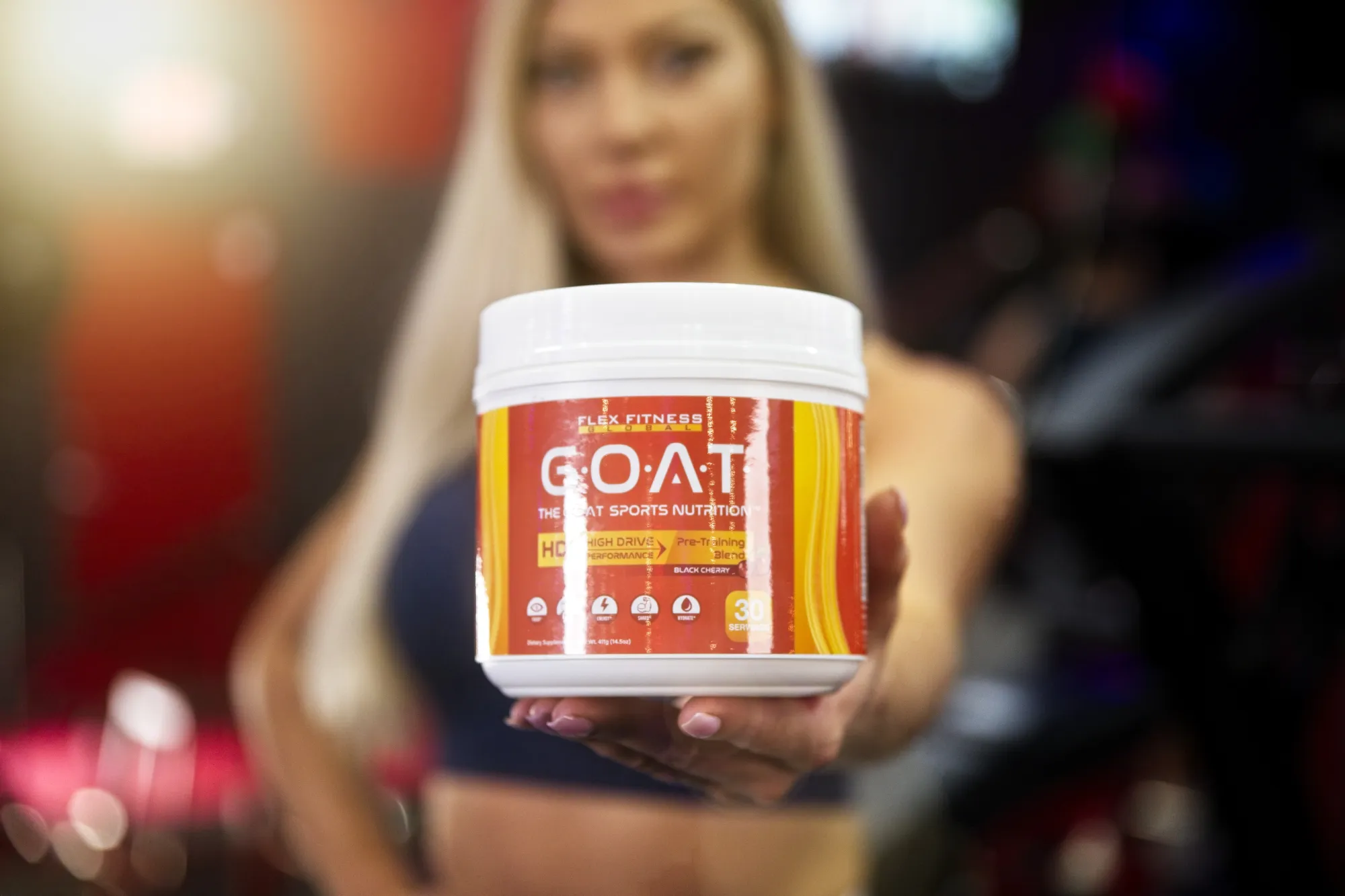 Woman holding a container of Flex Fitness GOAT sports nutrition supplement in a gym setting.