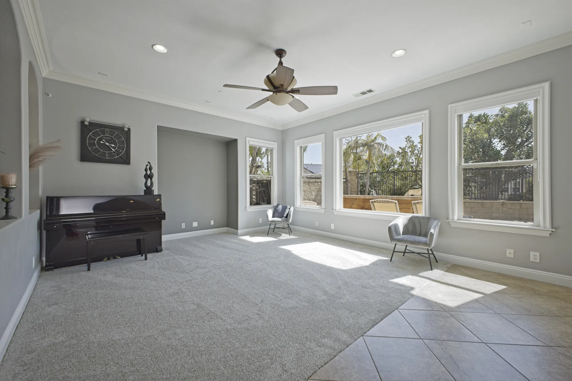 Bright living room with gray walls, carpeted floor, ceiling fan, upright piano, two gray chairs, and large windows showing outdoor greenery.